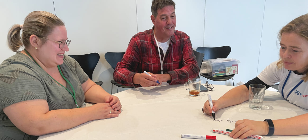 adviseur patiëntenparticipatie Martine Versluijs zit samen met 2 congresdeelnemers aan een tafel tijdens de tafellakensessie over richtlijnontwikkeling. Martine schrijft iets op met een stift. ets op tijdenstift iets op tijdens een tafellakensessie 