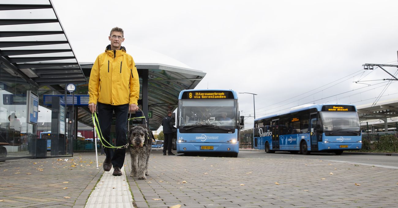 Man met hond loopt met geleidestok over geleidelijn op een stationsplein